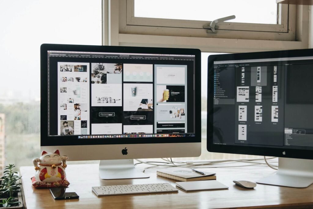 A modern workspace with two Apple iMac monitors displaying digital design software, showing various homepage layouts, UI components, and web design elements.