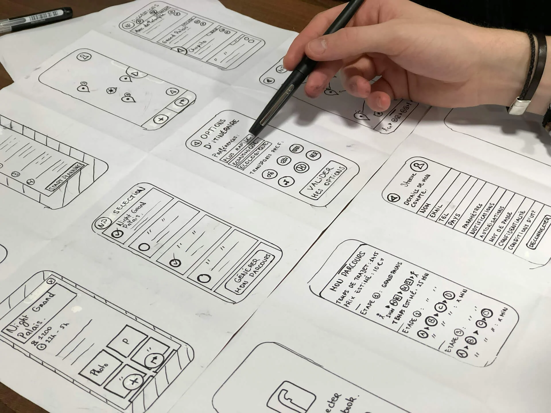 A person reviewing mobile app wireframes sketched on paper with a pen in hand, showing various screen layouts and UI elements.